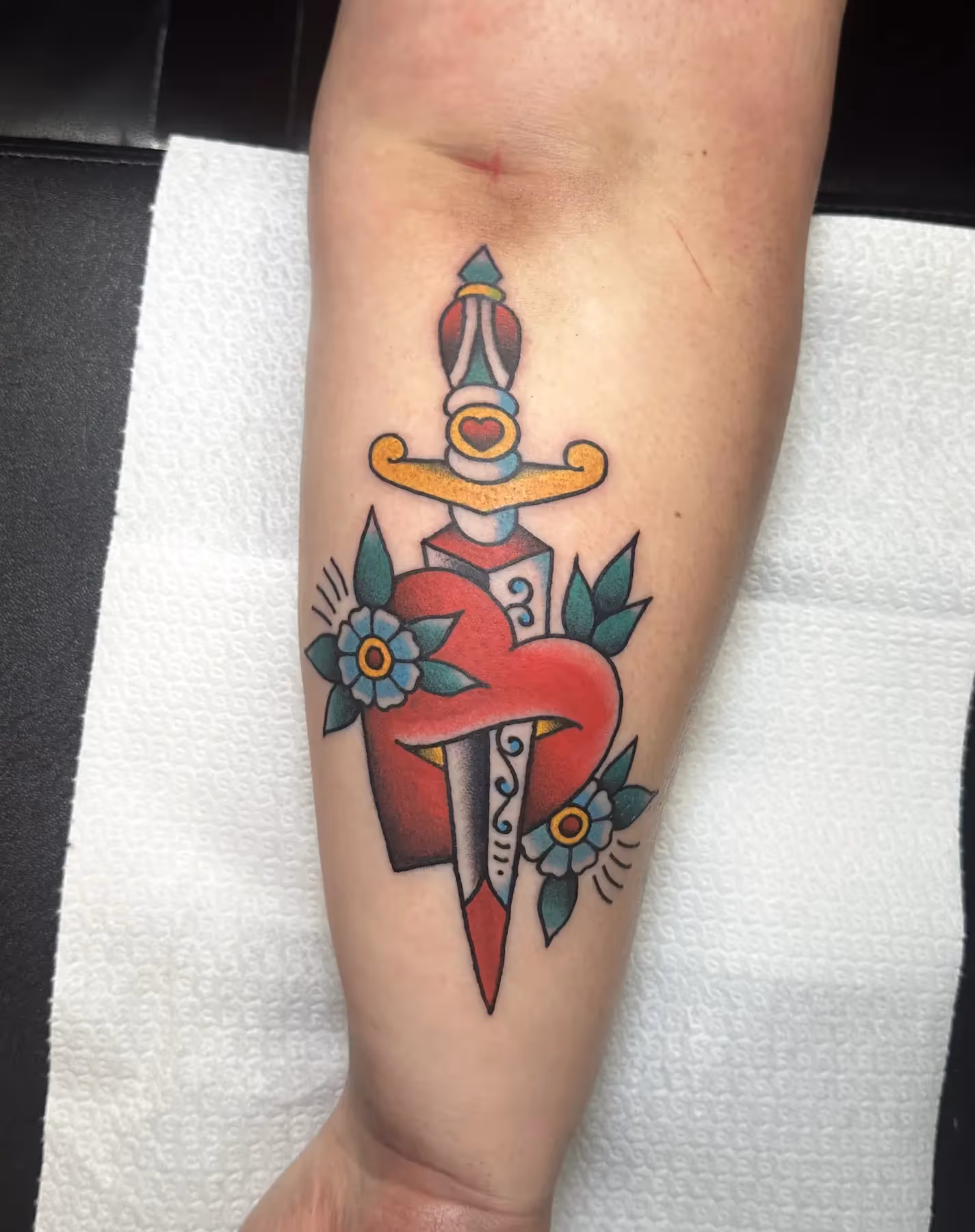 Traditional tattoo van een dolk door een hart met bloemen op onderarm, gezet door Nik Mart bij True Spirit Tattoo Barendrecht